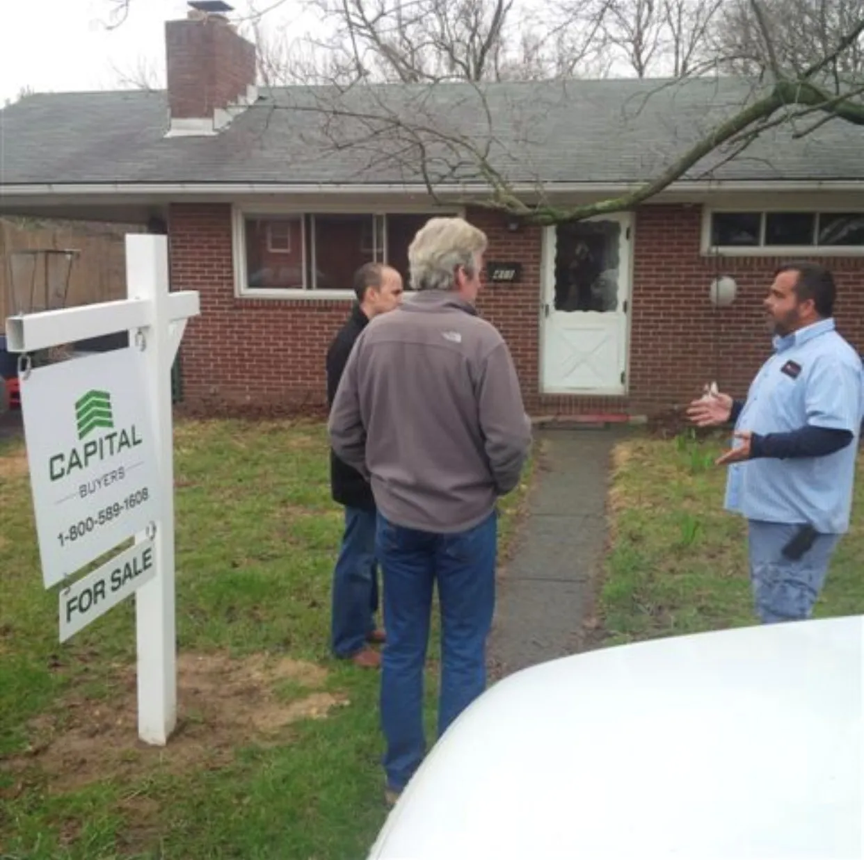 Three men discuss a property for sale in front of a brick house with a "For Sale" sign. The scene emphasizes real estate transactions and negotiations.
