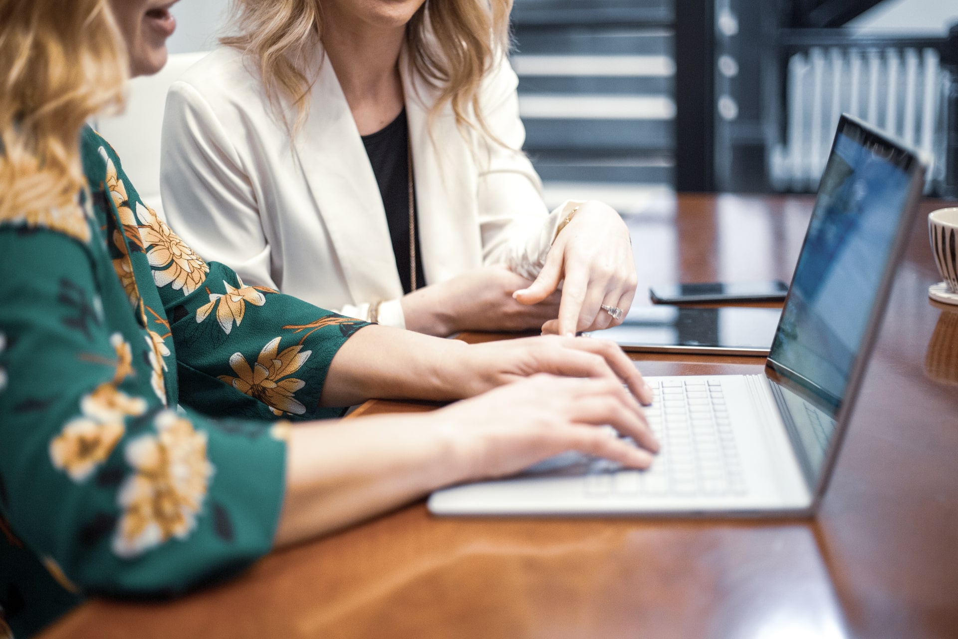 Two women collaborate at a desk, focusing on a laptop. One points while the other types, emphasizing teamwork and communication in a professional setting.