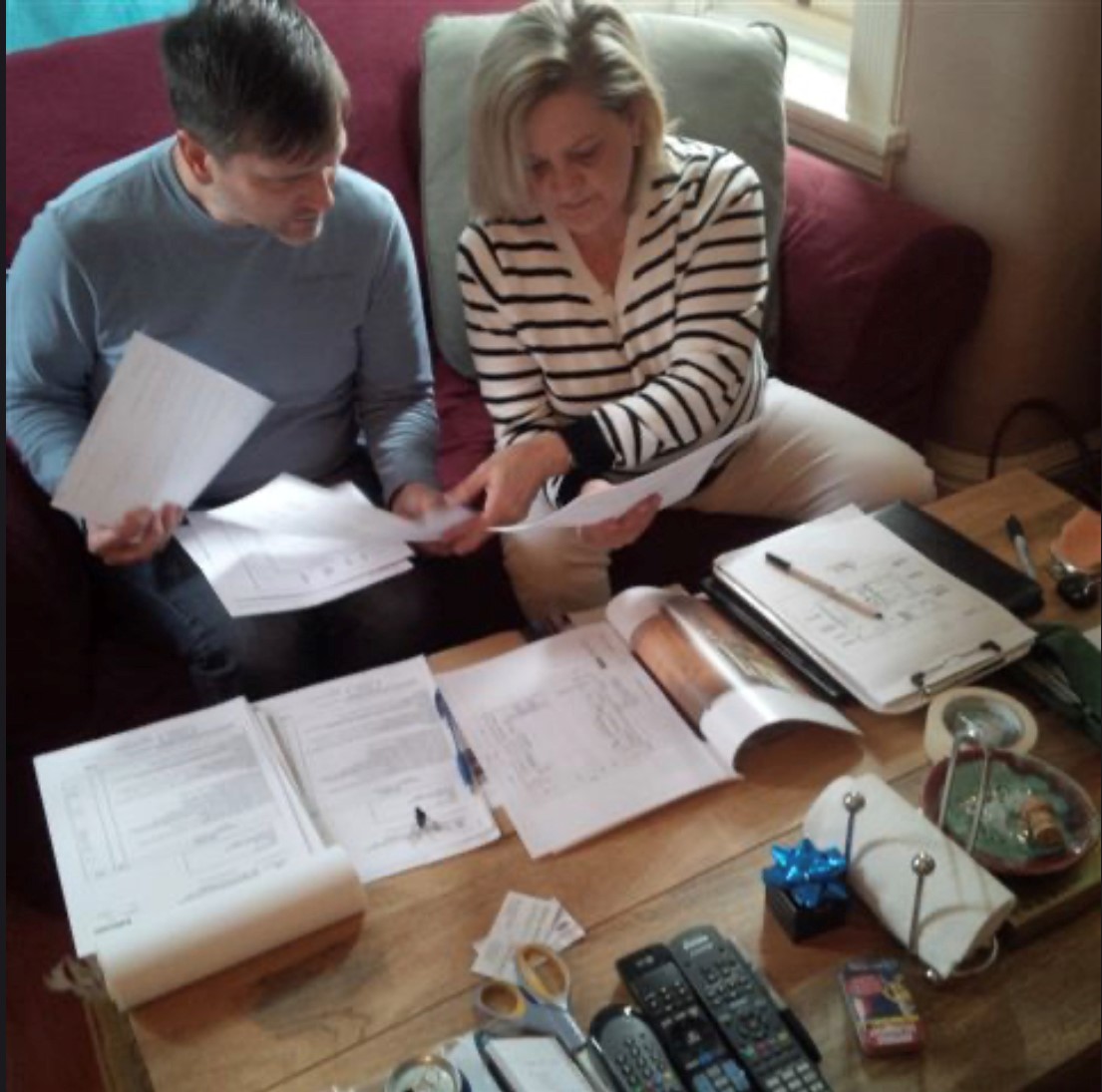 Two people review documents together on a coffee table scattered with papers, a notebook, and various household items, indicating a collaborative discussion.