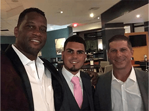 Three men pose together with smiles in a restaurant setting, showcasing a festive atmosphere. They're dressed in formal attire, indicating a special occasion.